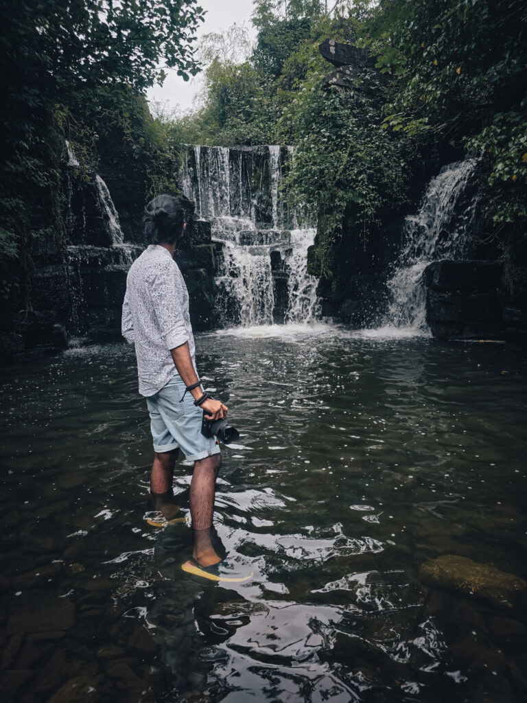 Hiker holding a camera beside a waterfall in Wales during a hiking and camping adventure.