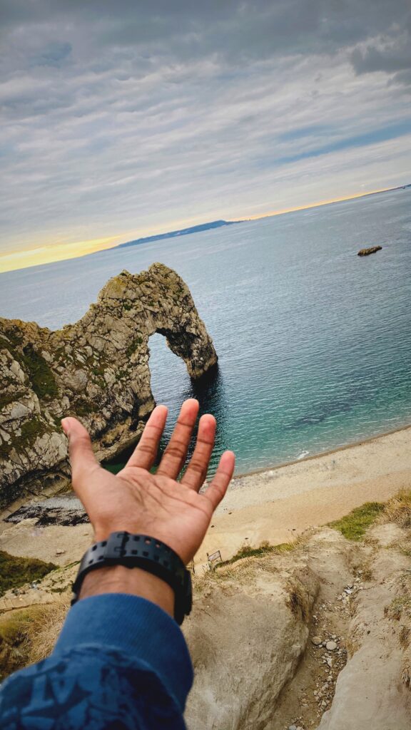 A hand gesturing toward the iconic Durdle Door rock arch overlooking the ocean in Dorset, UK